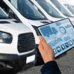 Hand holding tablet displaying fleet management data in front of white delivery vans, illustrating essential technology for Amazon DSP efficiency and safety compliance.