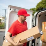 Delivery driver in red shirt and cap carrying a large package from a delivery van, representing Amazon Delivery Service Partner operations.